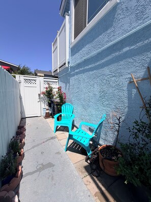 Ein Garten mit weißem Zaun, zwei türkisfarbenen Stühlen, einem Topfpflanzen und einer Holzleiter, die an einer blauen Wand anlehnt.