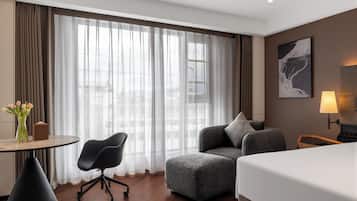 A hotel room with a bed, a desk with a chair, a sofa, a lamp, and a painting on the wall.