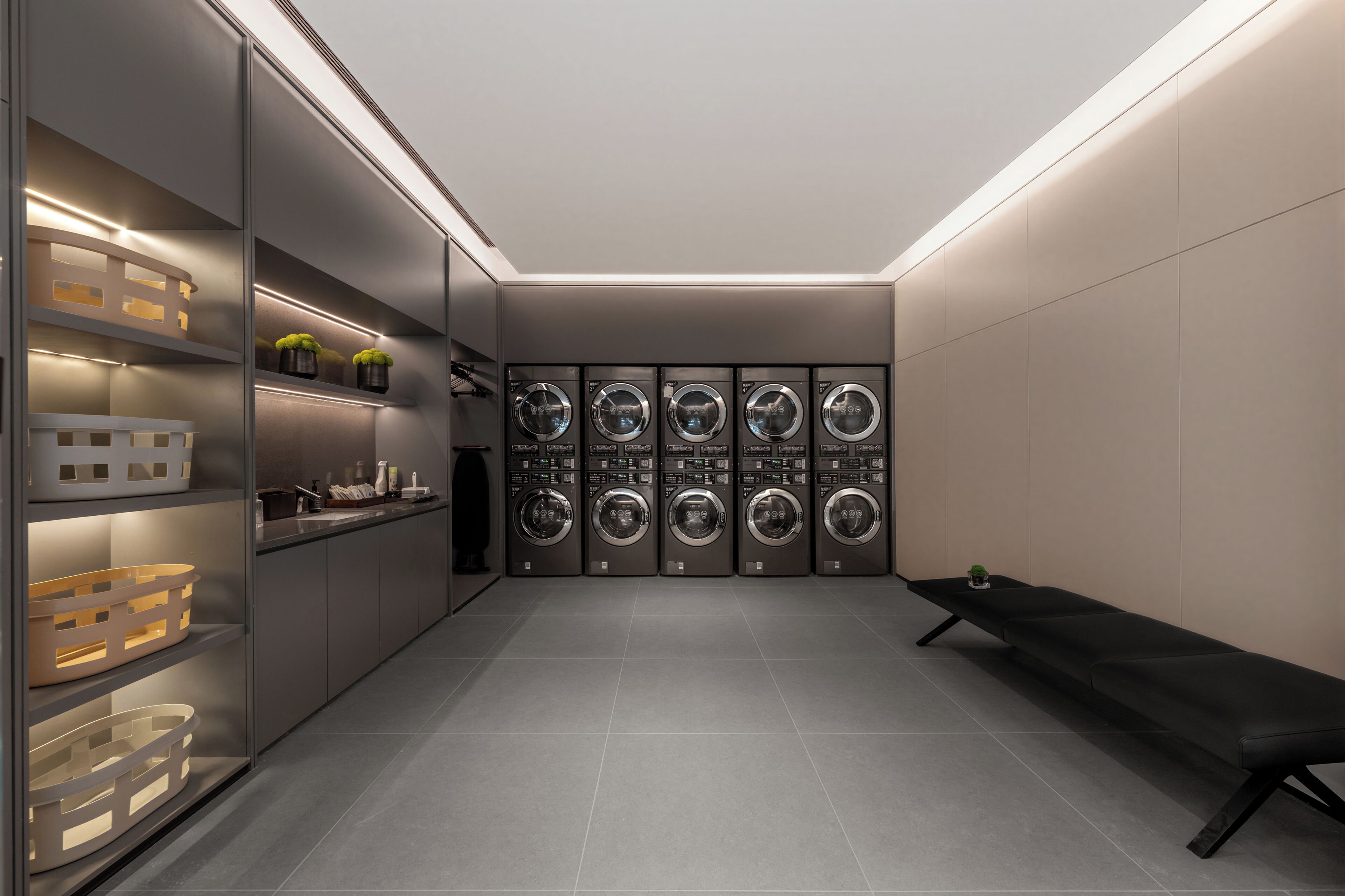 A modern laundry room with multiple washing machines, shelves with toiletries, and a bench.