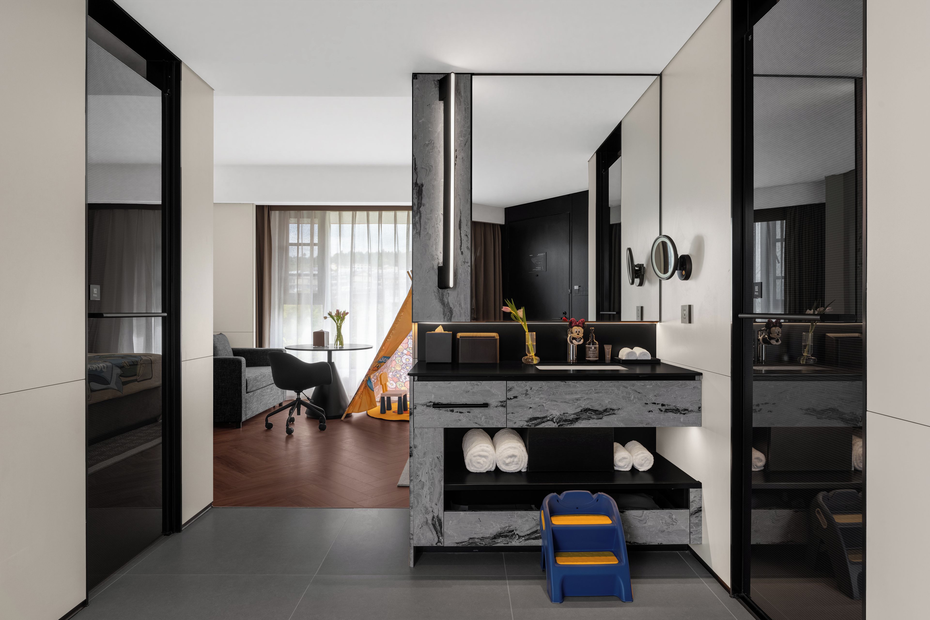 A modern hotel room with a marble fireplace, a sofa, a dining area, and a bathroom with a large mirror.