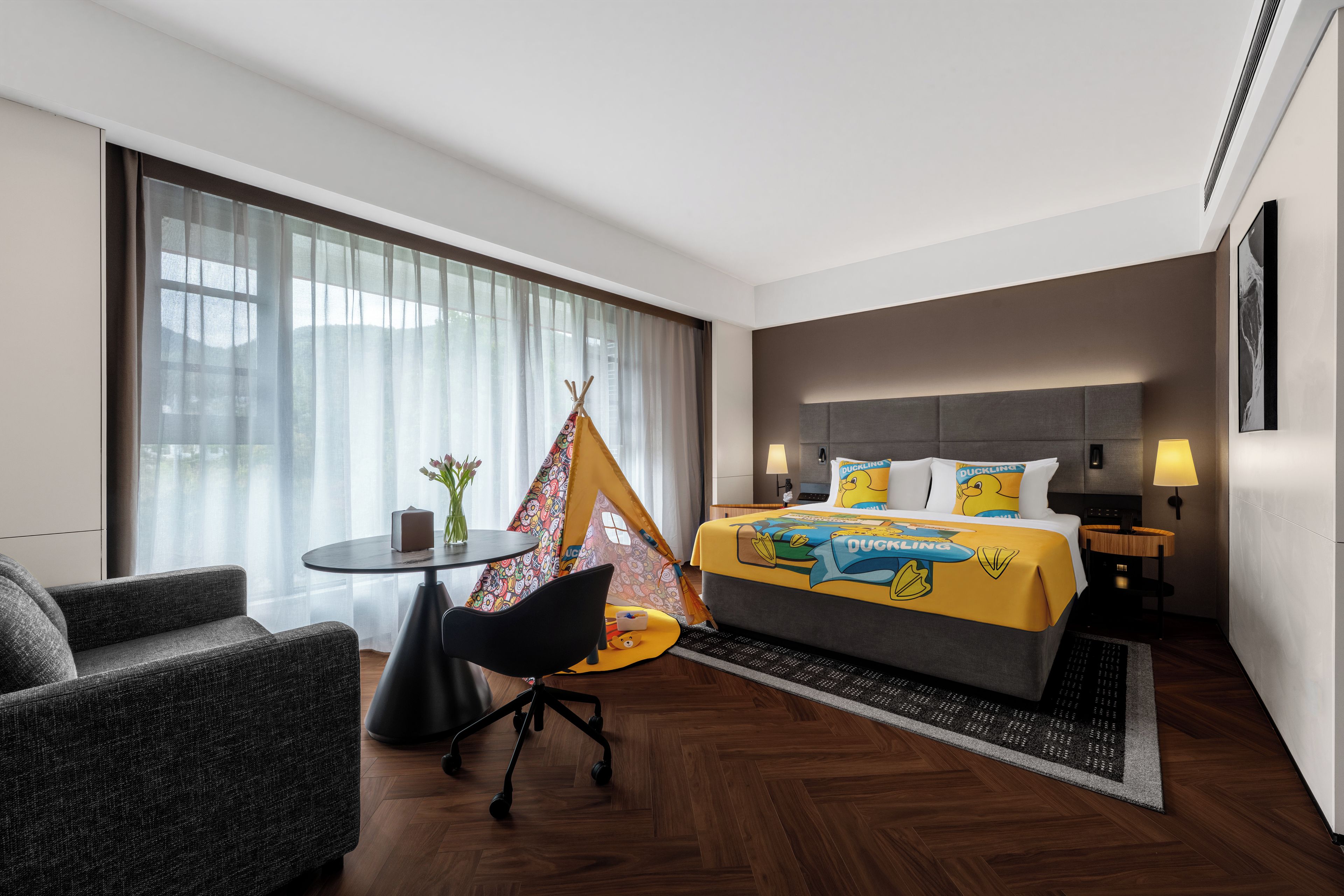 A hotel room with a bed, a small table, a chair, a window with curtains, and a wall-mounted lamp.