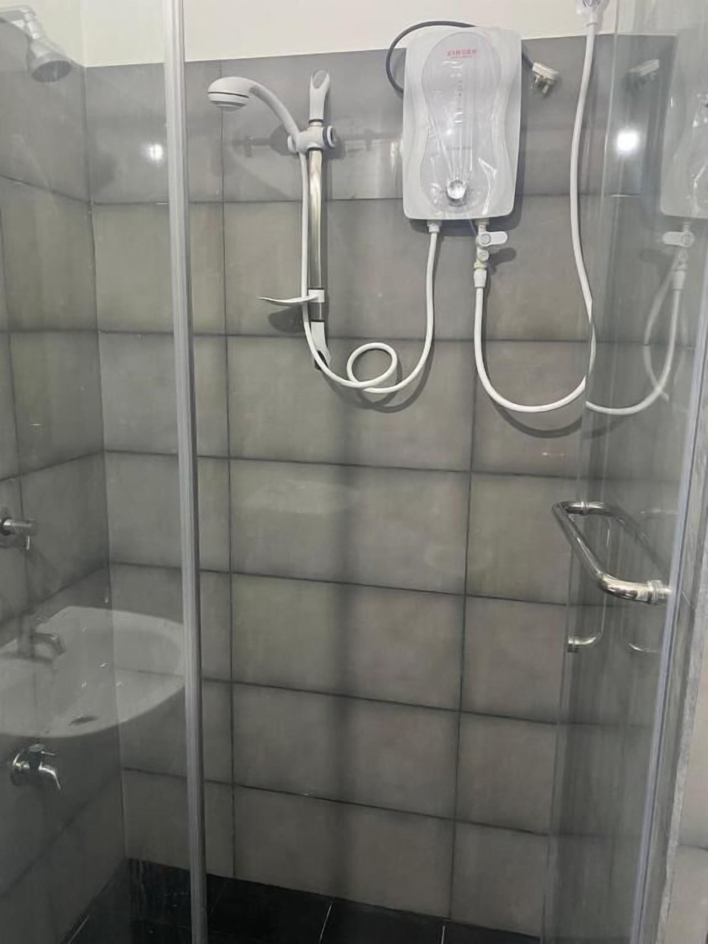 Chuveiro com chuveiro manual e torneira fixa na parede, pia com divisória de vidro e painel de controle do chuveiro.