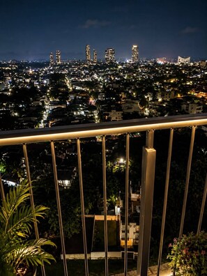 A night view of a cityscape with illuminated buildings and a balcony railing in the foreground.