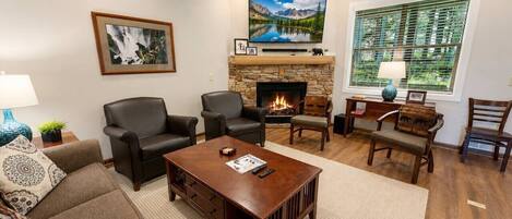 A living room with a fireplace, comfortable seating, a flat-screen TV, and a large window with blinds.