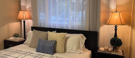 A neatly made bed with a checkered bedspread, two bedside lamps, and a window with sheer curtains.