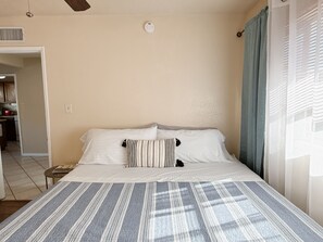 A bedroom with a bed, a bedside table, a window with curtains, and a view of a kitchen area. - Beautiful 3-bedroom house with big yard (Duarte)