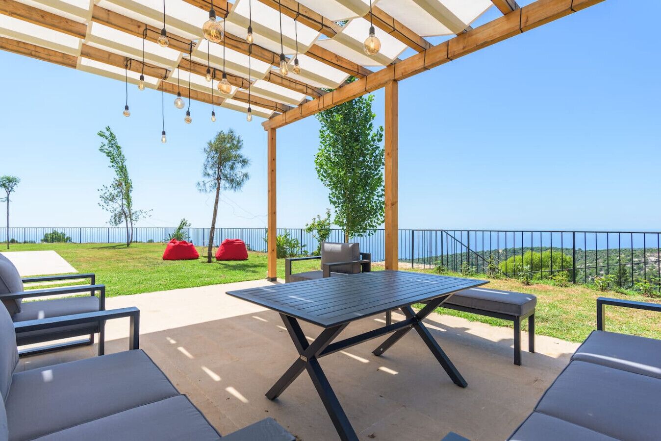 An outdoor patio with a table and chairs, a pergola with hanging lights, and a view of the sea.