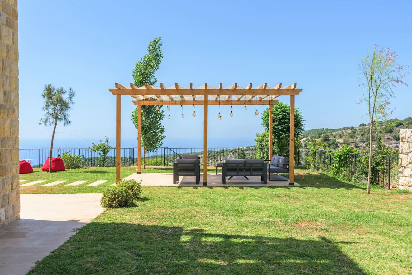 A wooden pergola with hanging lights, a seating area with a table and chairs, and a view of the sea.