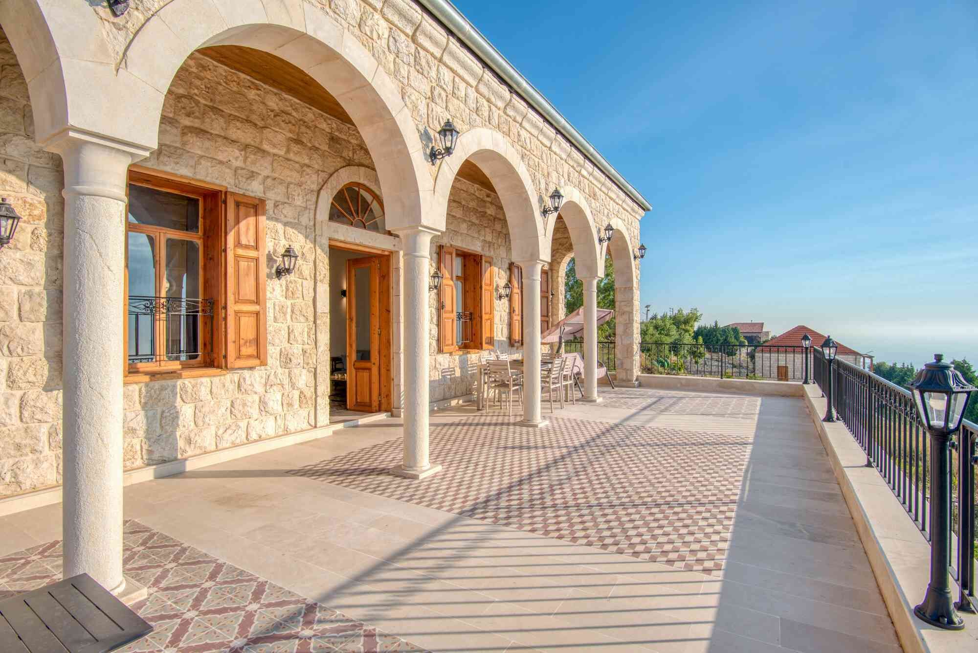 A stone building with arched windows and columns, a tiled patio, and a railing overlooking a landscape.