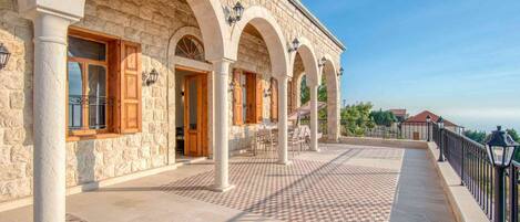 Un edificio de piedra con ventanas arqueadas y columnas, un patio con baldosas y una barandilla con vistas al paisaje.