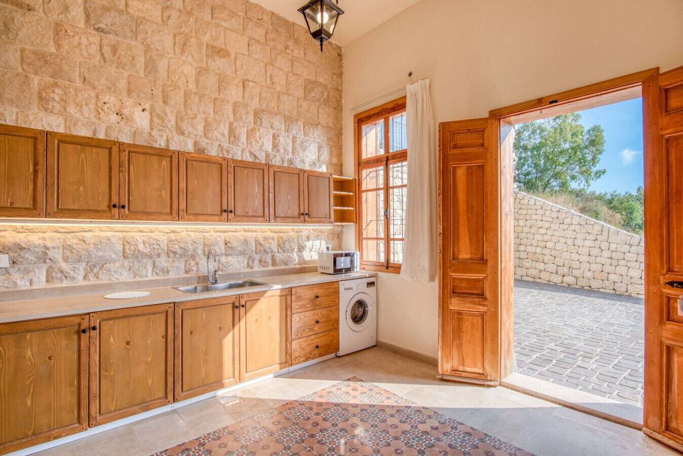 A kitchen with stone walls, wooden cabinets, a washing machine, and a view of a paved area outside.