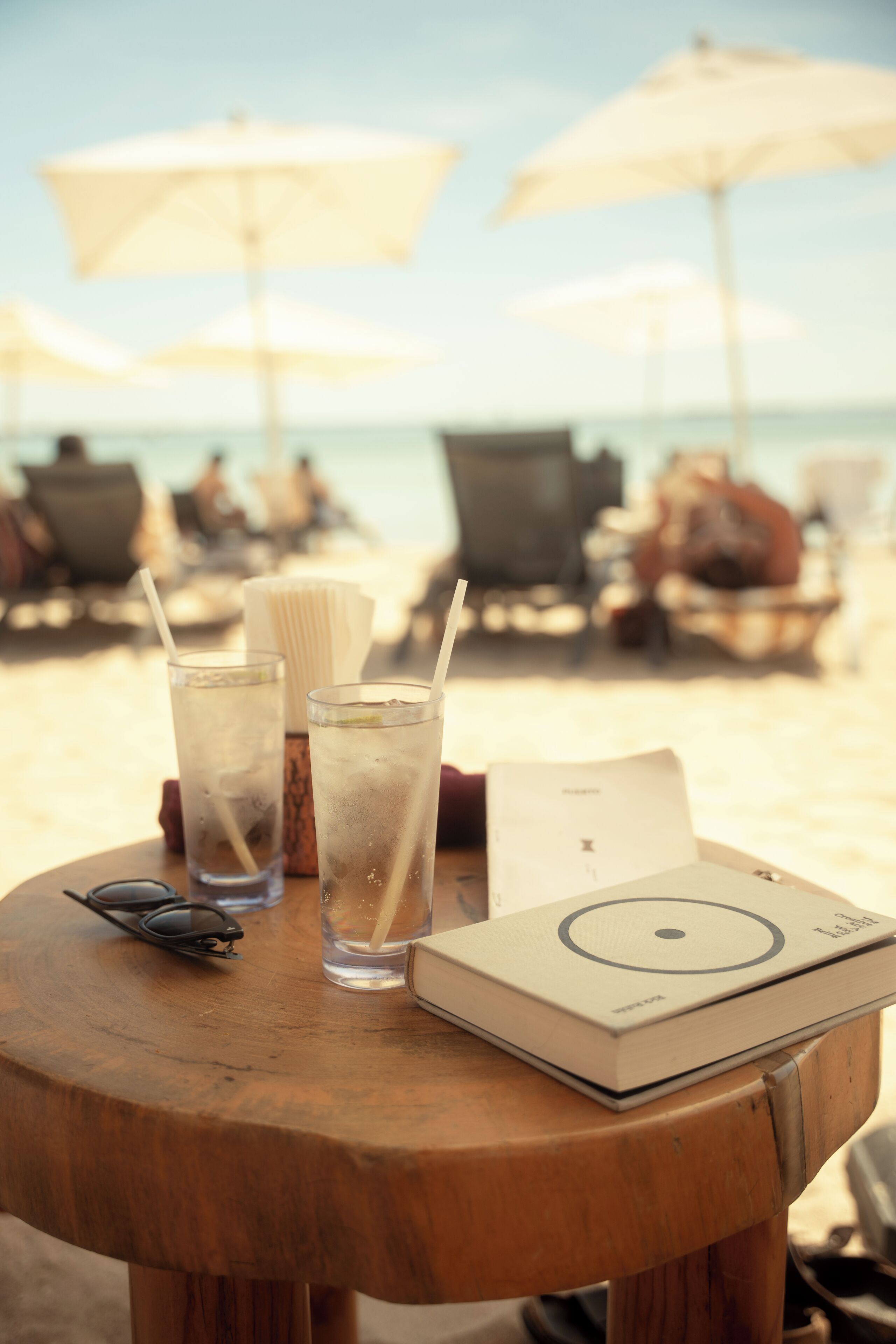 Une table ronde en bois, avec deux verres de boisson fraîche, une paire de lunettes de soleil et un livre.