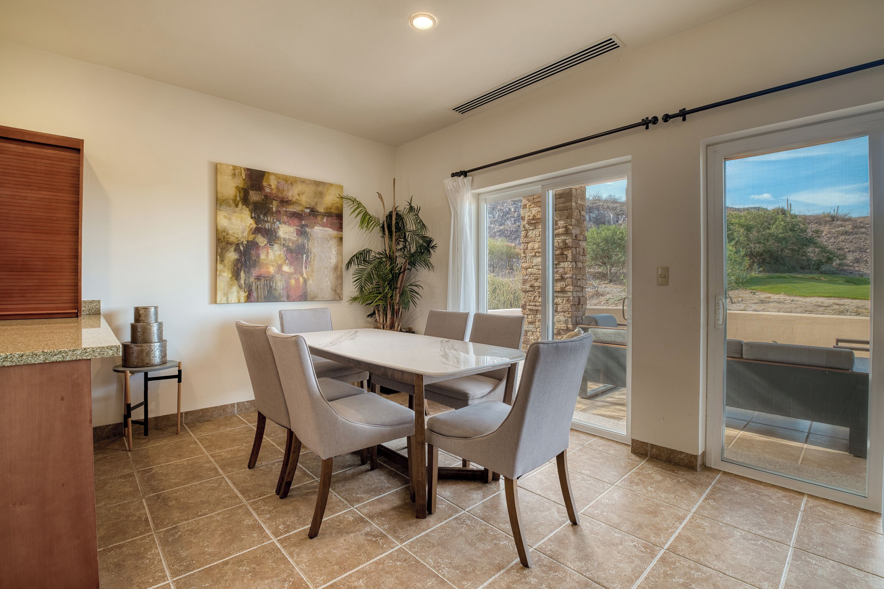 A dining area with a large table and chairs, a painting on the wall, and a view of the outdoors through a sliding glass door.