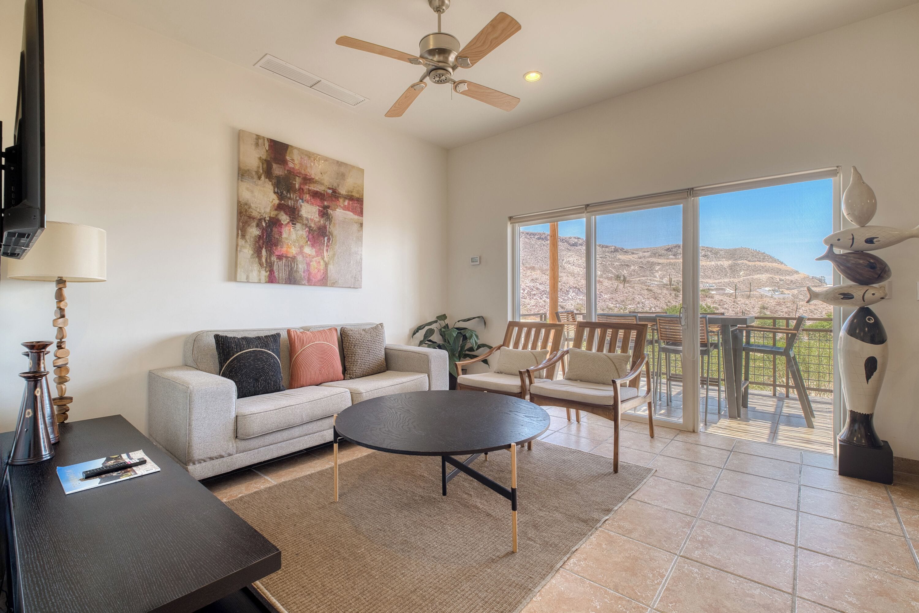 A spacious living room with a sofa, coffee table, and chairs, a ceiling fan, and a large window with a view of a mountainous landscape.