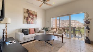 A spacious living room with a sofa, coffee table, and chairs, a ceiling fan, and a large window with a view of a mountainous landscape.