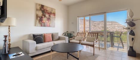 A spacious living room with a sofa, coffee table, and chairs, a ceiling fan, and a large window with a view of a mountainous landscape.