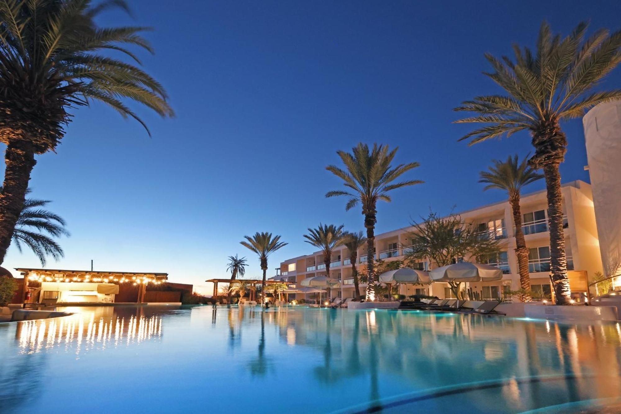 Zona de piscina hotelera con palmeras, sillones de descanso y un edificio de varios pisos.