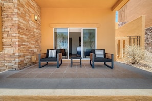 Un patio con dos sillas y una mesa, un muro de piedra y un ventanal corredizo que da a un espacio interior.