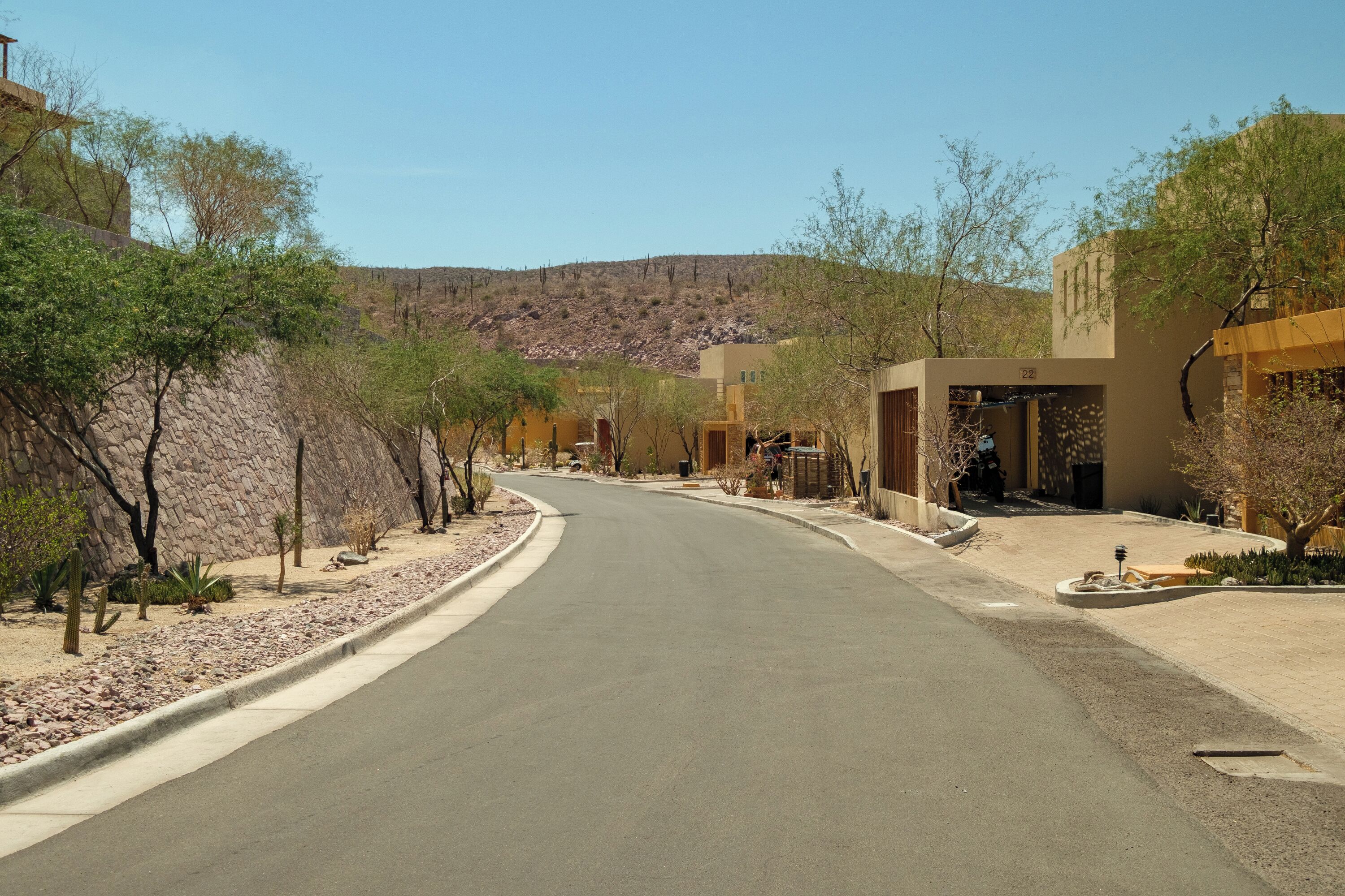 A curved road in a residential area with houses on both sides, a clear sky, and a hilly landscape in the background.