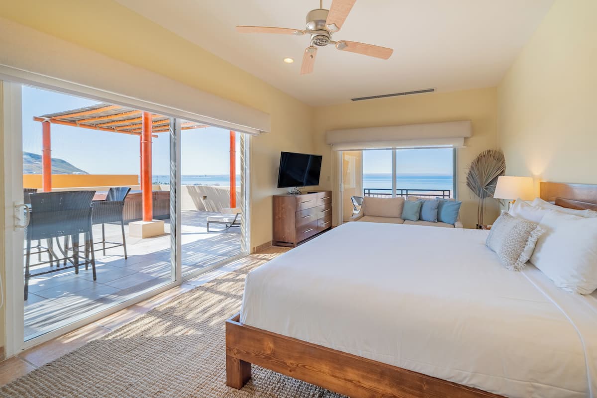 A bedroom with a large bed, a ceiling fan, a TV, and a view of the ocean.