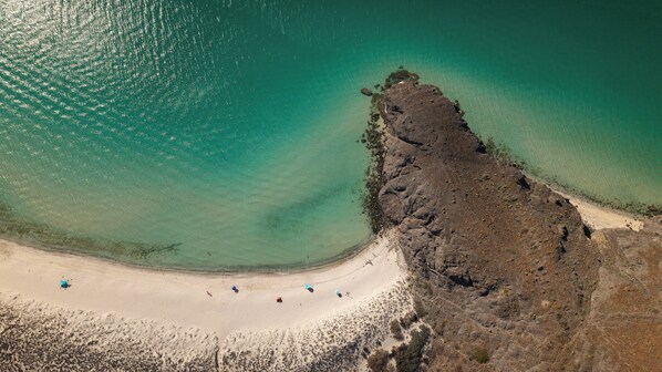 An aerial view of a coastal area with a sandy beach, clear turquoise waters, and rocky terrain.