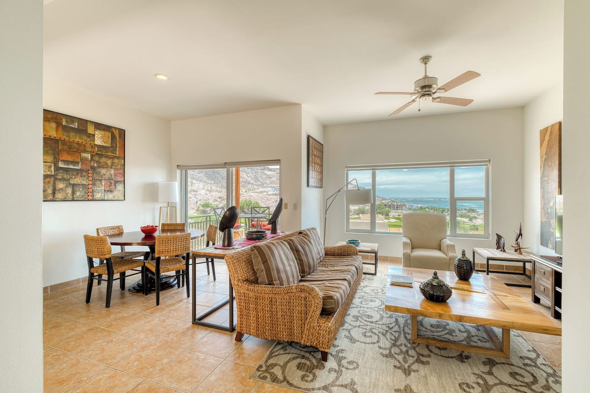 A spacious living room with a large sofa, dining table, and chairs. There is a ceiling fan, large windows with a view, and a rug on the floor.