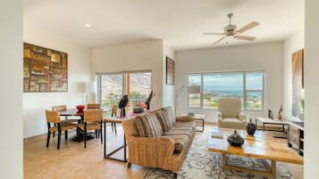 A spacious living room with a large sofa, dining table, and chairs. There is a ceiling fan, large windows with a view, and a rug on the floor.