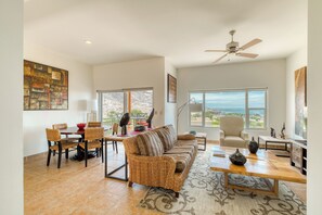 A spacious living room with a large sofa, dining table, and chairs. There is a ceiling fan, large windows with a view, and a rug on the floor.