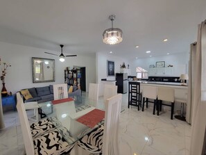 A modern living room with a glass dining table, a ceiling fan, and a kitchen area with white cabinets and a black countertop. - Villa Tropical - Penthouse 10 (Cabarete)
