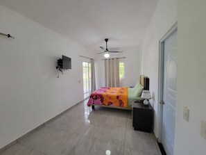 A bedroom with a bed, a nightstand, a TV mounted on the wall, a ceiling fan, and a door. - Villa Tropical - Penthouse 10 (Cabarete)