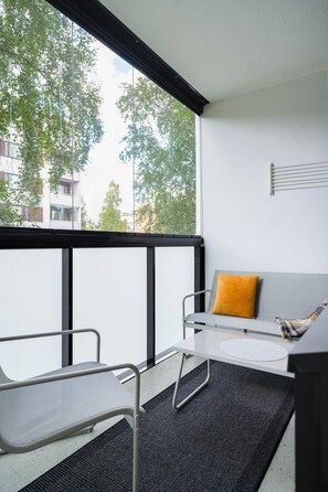 A balcony with a table and chairs, a sofa with a cushion, and a view of trees and buildings. - Pondside City Flat By Lapland Host (Rovaniemi)