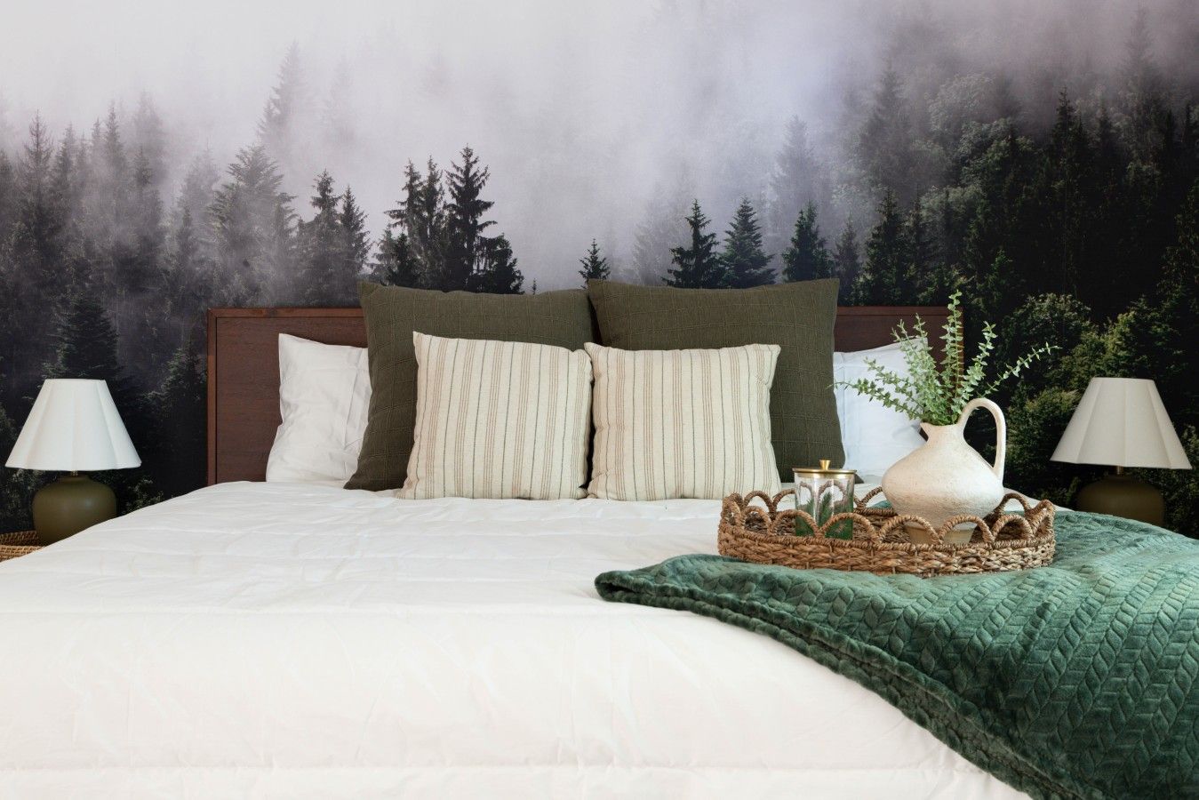 A bed with a white duvet, two pillows, a green throw, and a woven basket with a pitcher.
