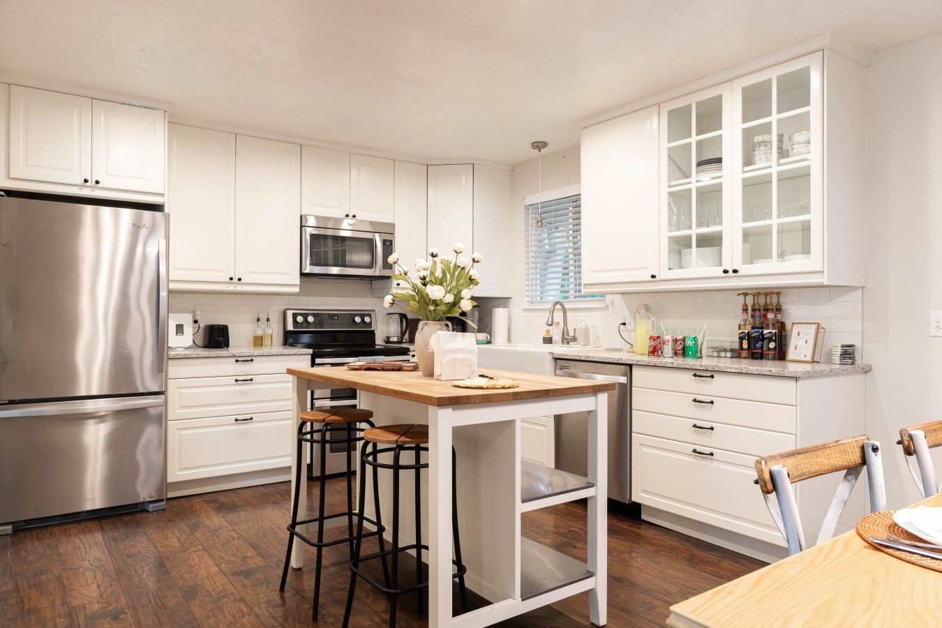 A modern kitchen with white cabinetry, stainless steel appliances, a central island with stools, and a dining area with a wooden table.