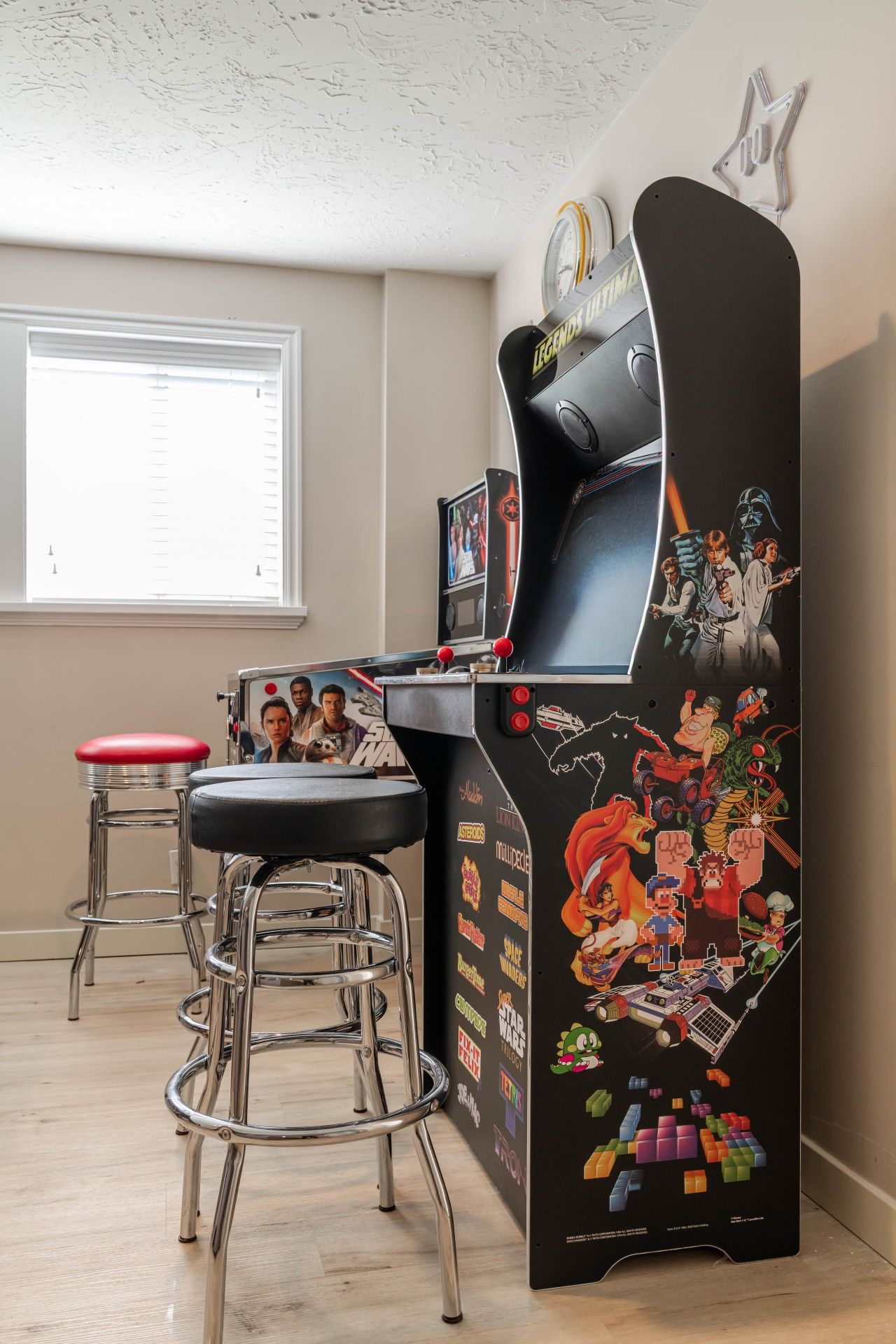 Ett rum med ett spelarkad, två pallar, ett fönster med persienner och en vägg full med olika klistermärken.