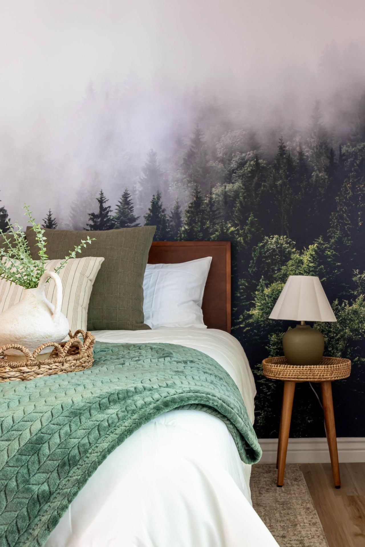 Un dormitorio con cama, almohadas, una mesita de noche con lámpara y un mural en la pared con un bosque.