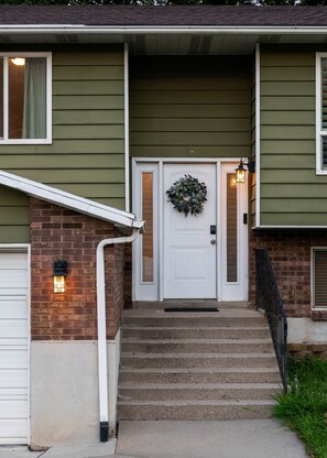 A two-story house with a front door, a brick wall, a white door with a wreath, and a small staircase leading to the entrance. - Hot Tub+Game Room, 8 Ski resorts + SLC | Sleeps 14 (Centerville)