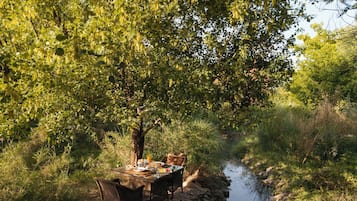 An outdoor dining area with a table and chairs set up on a stone ledge by a stream, surrounded by lush greenery.