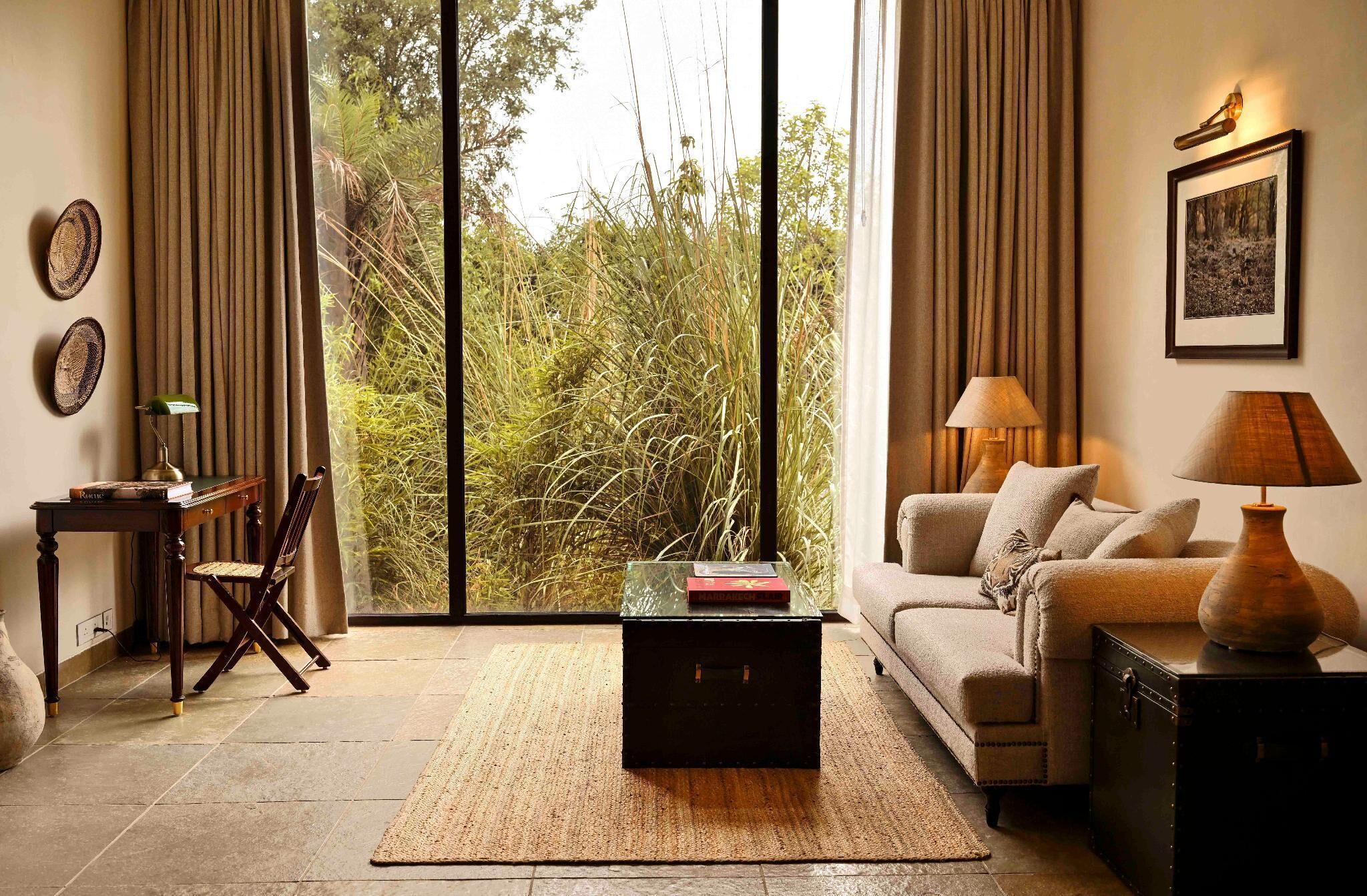 A living room with a sofa, a coffee table, a lamp, and a view of greenery through large windows.