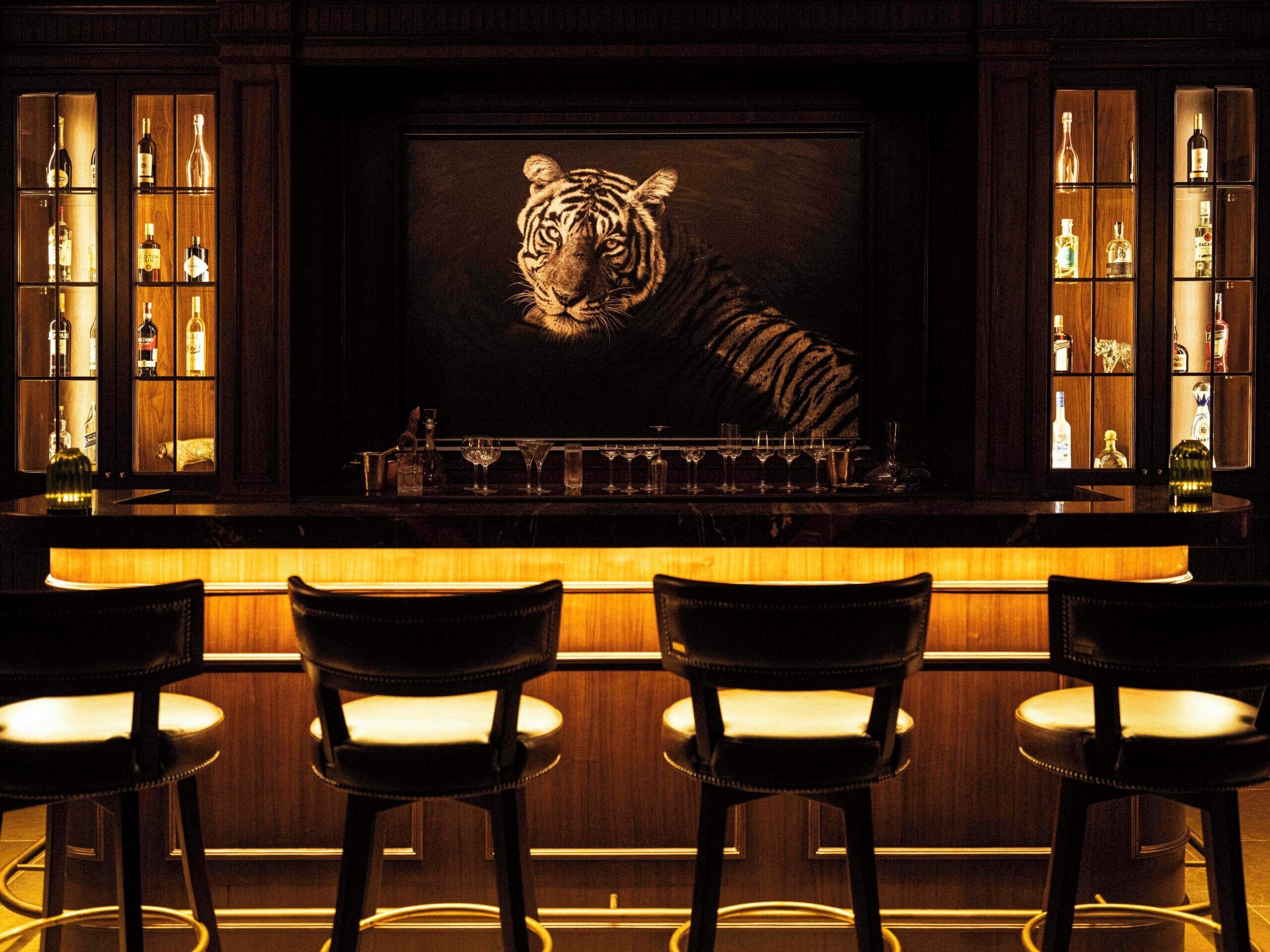 Zona de bar con un mural de tigre, estantes de madera con botellas y tres taburetes.