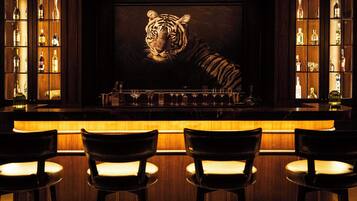 Zona de bar con un mural de tigre, estantes de madera con botellas y tres taburetes.