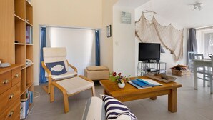 Una sala de estar con una estantería de madera, un sillón acolchado con un cojín con forma de pez, una mesa de centro con un jarrón de flores, un televisor sobre un mueble bajo y un gran tapiz tejido en la pared.