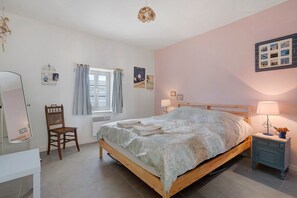 A bedroom with a wooden bed, a chair, a bedside table with a lamp, and a window with curtains.