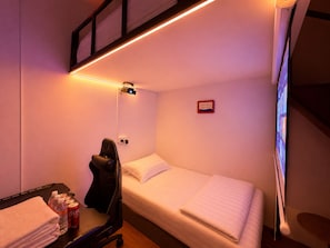 A single bed with white bedding, a desk with a chair, a bottle of water, a can of Coca-Cola, and a projector mounted on the wall.
