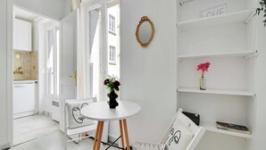 Et lyst interiør med et rundt bord, en stol med ansigtspude, en reol med en vase med blomster og et spejl på væggen.