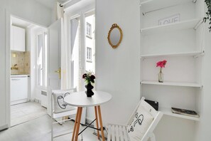 Ein helles Interieur mit einem runden Tisch, einem Sessel mit Gesichtsauflage, einer Kommode mit Blumenvase und einem Spiegel an der Wand.
