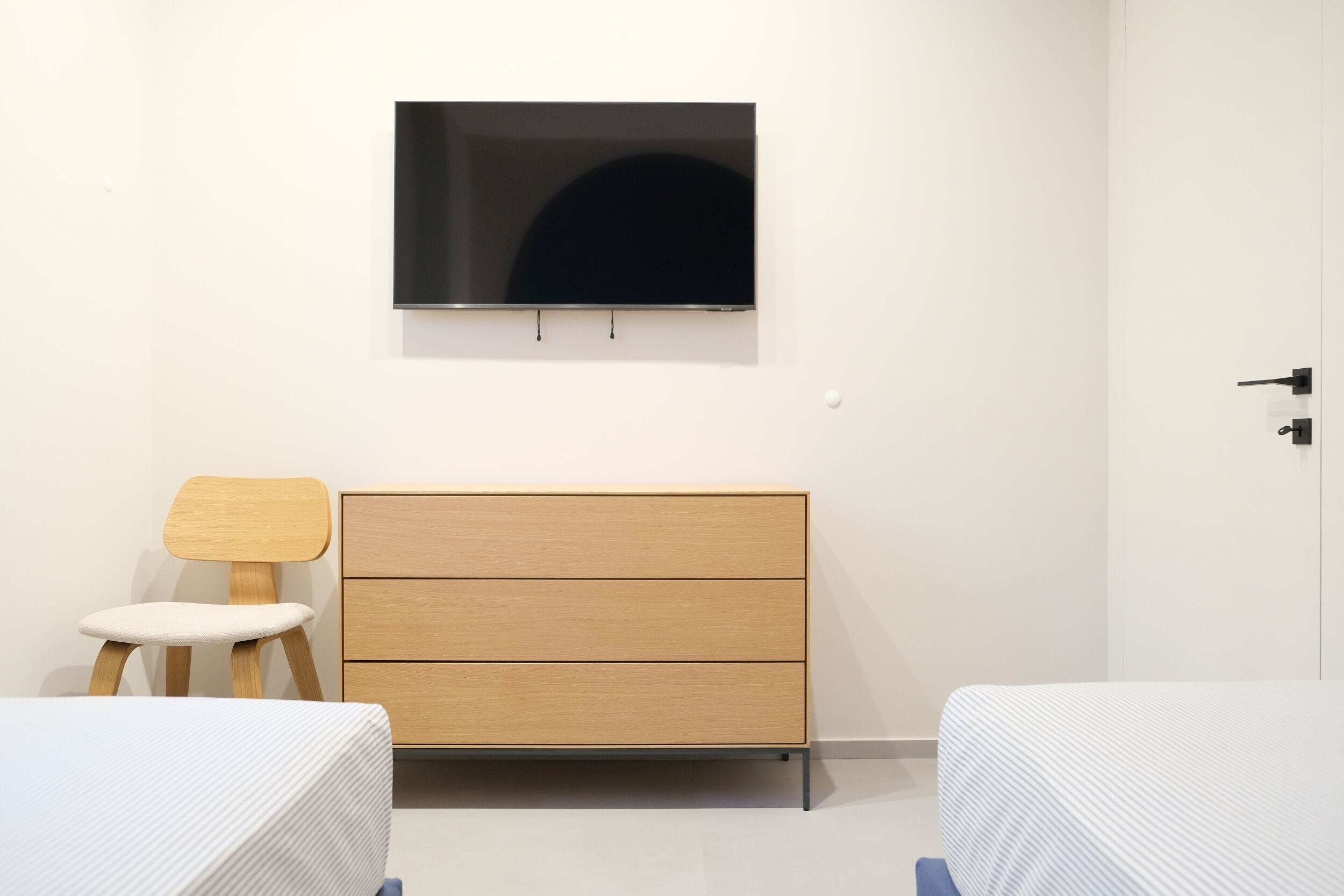 Una habitación minimalista con una cama, un tocador de madera, una silla y un televisor en la pared.