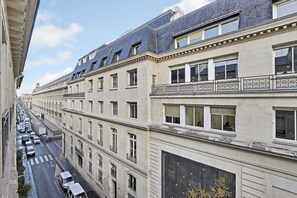 A multi-story building with a row of windows on the ground floor, a balcony with decorative railings, and a street with parked cars below. - Charming studio 2P - Musée d'Orsay / Louvre (Paris)