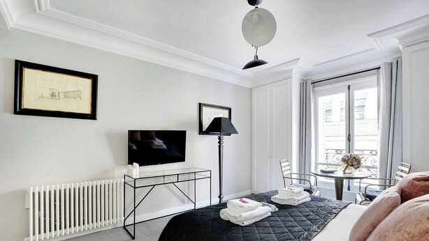 A hotel room with a bed, a desk with a flat-screen TV, a radiator, a window with a view, and a chair. - Charming studio 2P - Musée d'Orsay / Louvre (Paris)