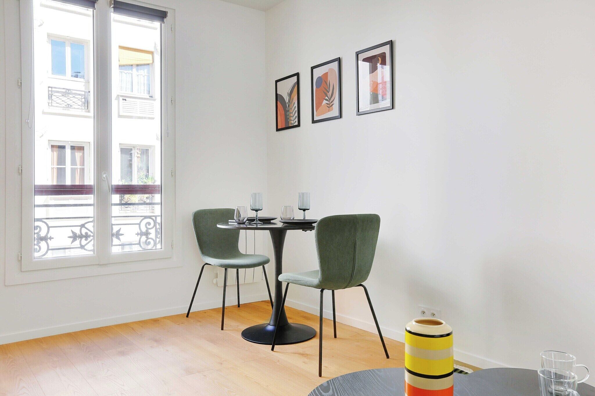 A room with a dining table, two chairs, a window with a view of buildings, and three framed pictures on the wall.