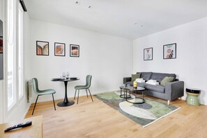 A modern living room with a grey sofa, two green chairs, a round dining table, and a coffee table. There are framed artworks on the walls and a remote control on the floor. - Charming studio for 2P - near Parc Monceau (Paris)